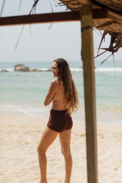 Sienna mini shorts in deep brown, high-waisted with smooth stretch fabric, shown on model from side view on sandy beach.
